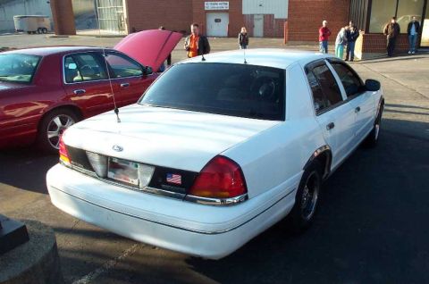 1998 Ford Crown Victoria - A white 1998 Ford Crown Victoria is parked next to a red car with its hood open, with several people standing around.