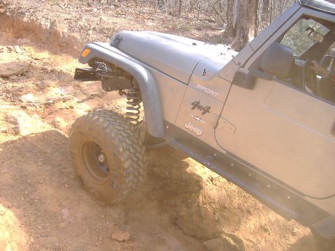 2004 Jeep Wrangler - A tan Jeep Wrangler 4x4 Sport is shown tackling a challenging dirt trail, with its front tire angled on an incline.