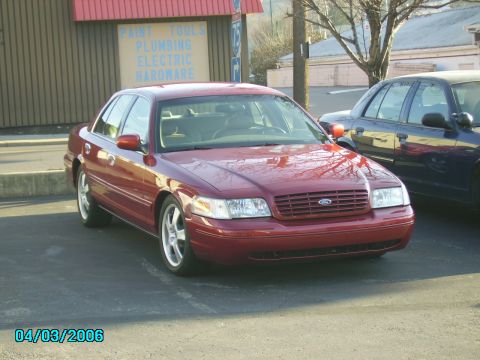 1998 Ford Crown Victoria - A red 1998 Ford Crown Victoria is parked in front of a building with visible text. Another vehicle is parked to its right.