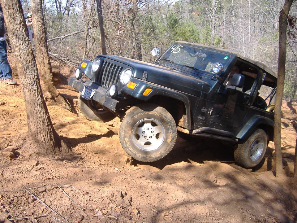 Dark green Jeep TJ climbing a steep, dirt trail with trees on either side. A person stands near a tree on the left