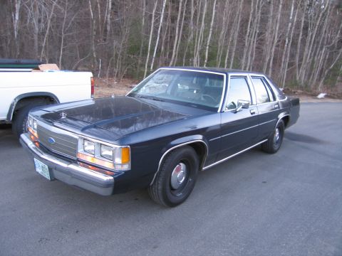 1991 Ford LTD Crown Victoria - A dark blue 1991 Ford Crown Victoria is parked on asphalt with a white pickup truck to its left and trees in the background.