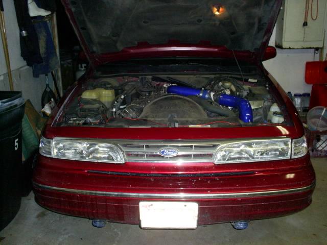 Front of red Ford sedan, hood open, blue intake piping and Ford grille badge
