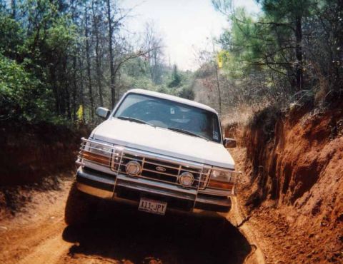 1996 Ford Bronco - A 1996 Ford Bronco navigates a challenging off-road trail surrounded by trees and dry brush.