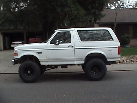 1994 Ford Bronco - A white, lifted SUV is parked on the side of a residential street, showing its side profile and large off-road tires.
