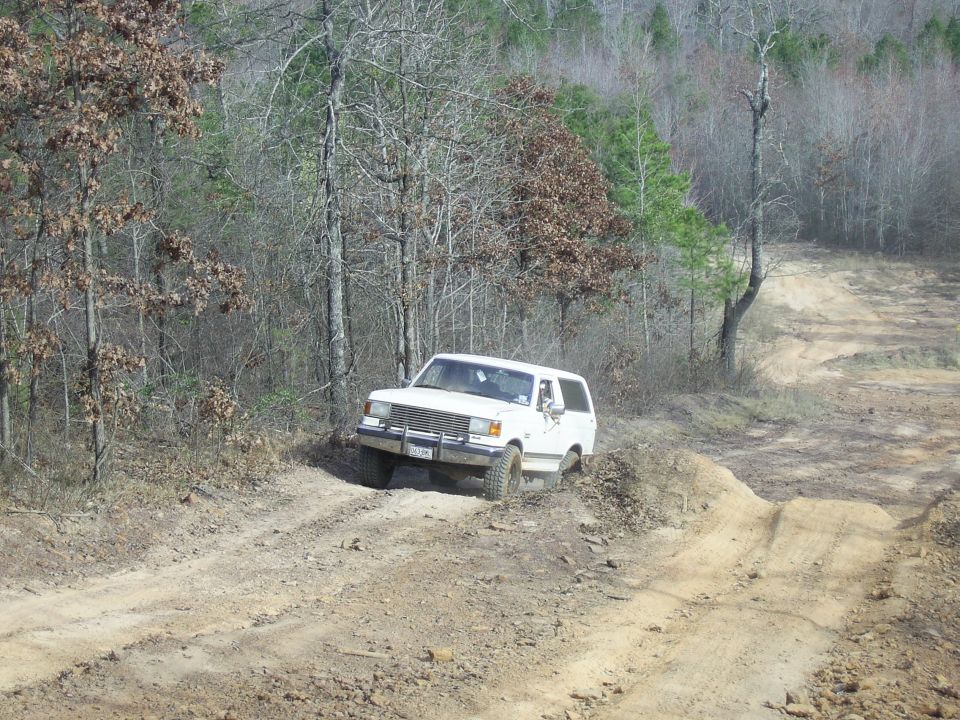 White Ford Bronco with brush guard climbing a rutted clay trail in bare winter woods