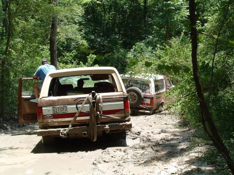 1993 Ford Bronco - Two Ford Broncos navigate a muddy trail, with one vehicle partially submerged in the mire.