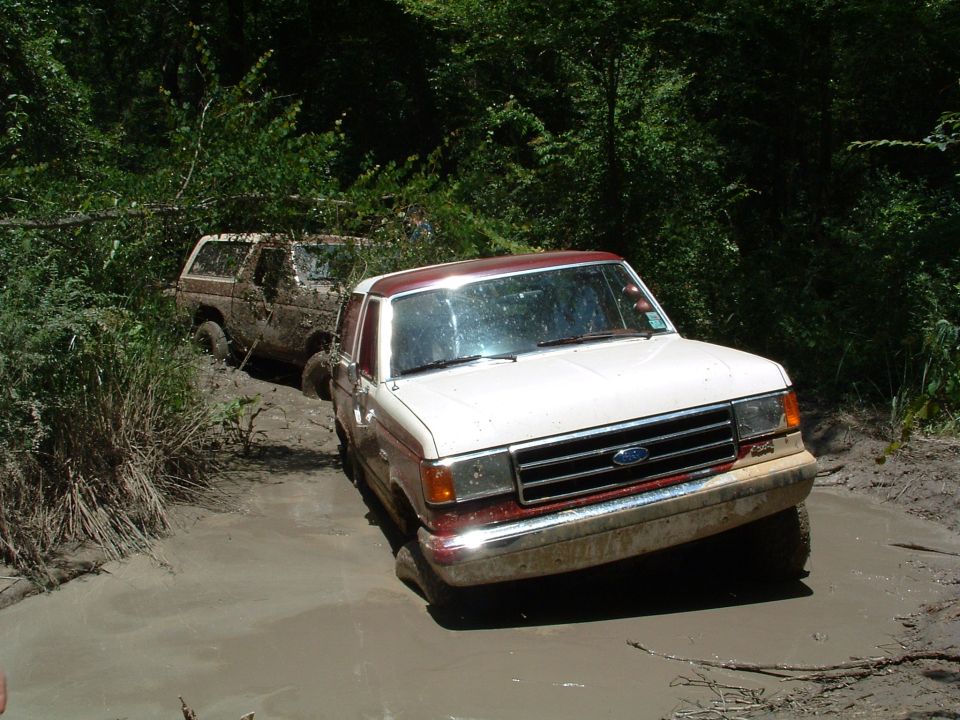 Two Ford trucks stuck in mud, one in foreground with white and red paint, other behind it covered in mud
