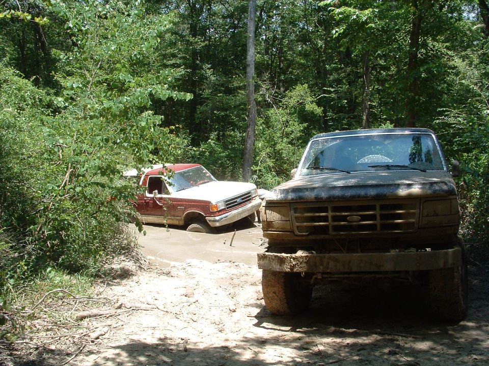 Mud-covered Ford F-Series truck front view, with another maroon and white Ford truck stuck in mud behind it