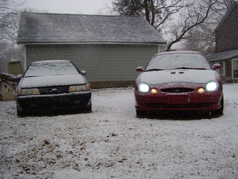 1997 Ford Taurus - Two Ford Taurus sedans are parked in a snowy yard in front of a garage.