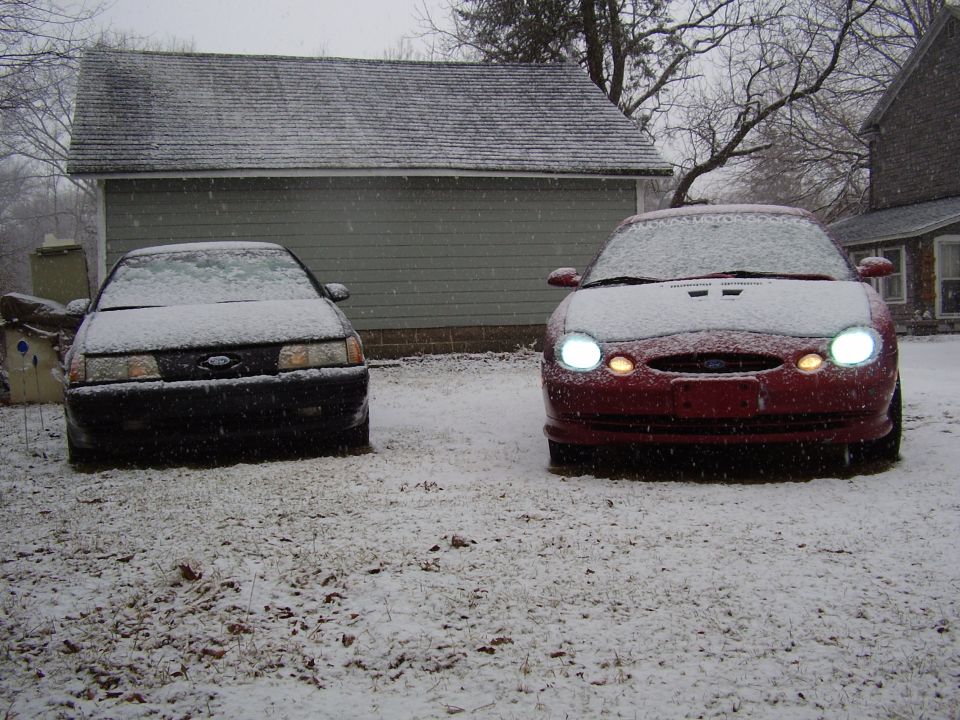 Two Ford Taurus sedans covered in snow, parked in front of a garage