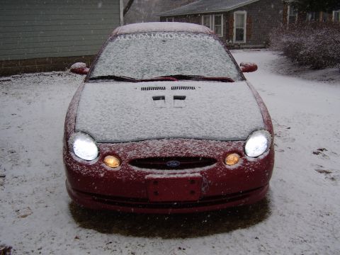 1997 Ford Taurus - A red 1997 Ford Taurus sits covered in snow, its headlights illuminating the winter scene.