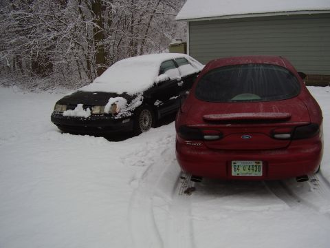 1997 Ford Taurus - Two sedans sit parked in a snowy driveway next to a garage.