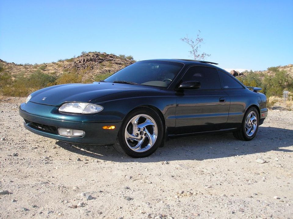 Dark green 1993 Mazda MX-6 coupe with chrome wheels parked on a dirt road
