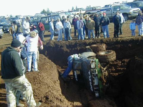 1994 Ford Explorer - A green off-road vehicle is stuck on its side in a deep mud pit, surrounded by onlookers.