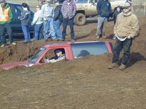 1994 Ford Explorer - A red SUV is deeply mired in mud, with onlookers observing the scene from the embankment.