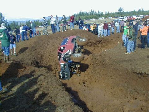 1994 Ford Explorer - A red 1994 Ford Explorer is overturned in a mud pit during an event, with onlookers gathered around.