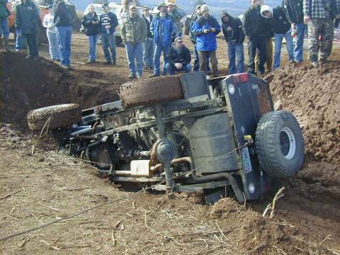 1994 Ford Explorer - An overturned black SUV is stuck deep in mud, with onlookers gathered around.