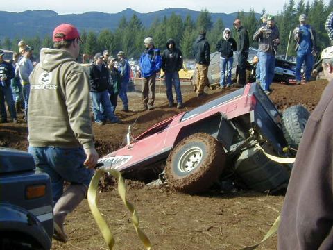 1994 Ford Explorer - A red off-road vehicle is stuck in a muddy pit, with a crowd of onlookers observing the scene.