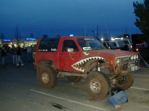 1994 Ford Explorer - A person works on a lifted red SUV with shark teeth graphics, parked in a lot at dusk.