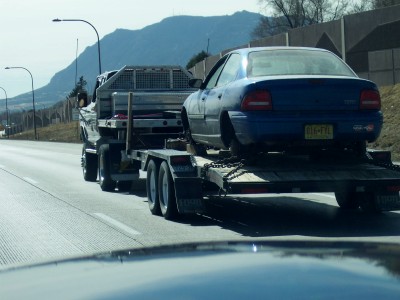 1972 Ford Ranchero - A blue sedan is transported on a trailer behind a truck on the highway.