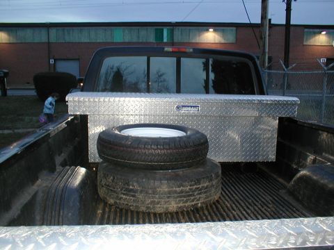 1979 Ford Bronco - The bed of a pickup truck holds a toolbox and spare tires, with a building and a person in the background.