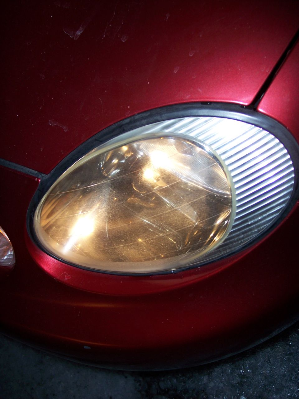 Red car headlight with visible scratches on the lens, part of the front fender visible