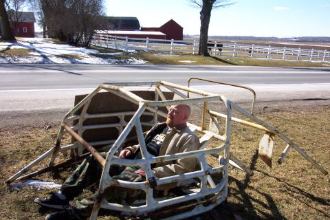 1979 Ford F-150 - A man sits in a rusty white roll cage in a rural setting with farm buildings in the background.