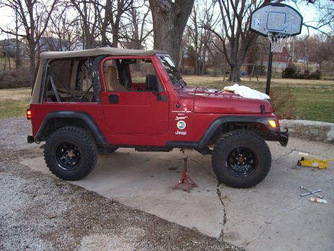 1998 Jeep TJ - A red Jeep Wrangler is shown with a tan soft top and lifted suspension, parked on a cracked concrete surface.