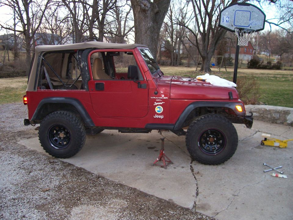 Red Jeep Wrangler with a tan soft top, lifted suspension, and large tires, parked on a cracked concrete driveway