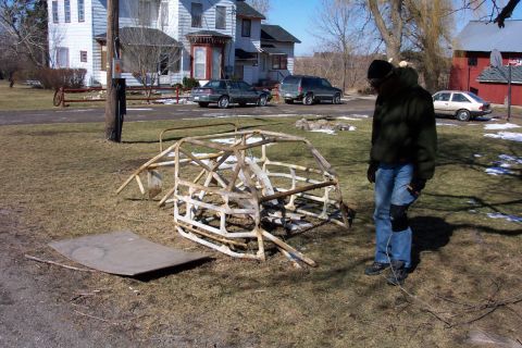 1979 Ford F-150 - A rusted car frame sits in a yard with other vehicles and houses visible in the background.