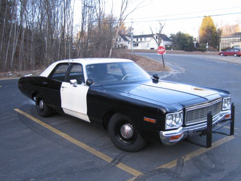 1991 Ford LTD Crown Victoria - black-and-white police-style Dodge sedan parked at an angle in a lot, with leafless trees and a stop sign in the background.