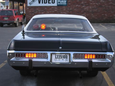 1991 Ford LTD Crown Victoria - Rear view of a black police sedan showing lit taillights and a glowing amber rotating beacon, parked before a brick wall.