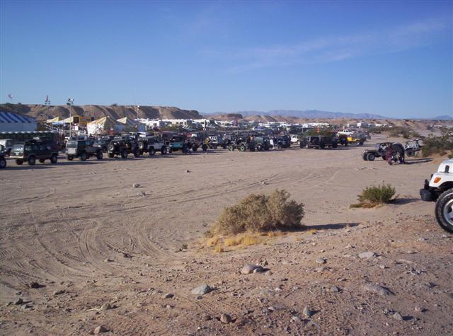 many off-road vehicles parked in a dusty lot with tents and hills in background