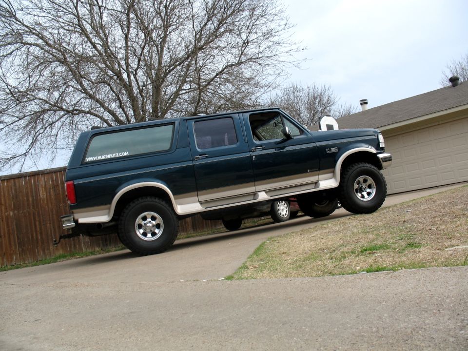 Green Ford Bronco full-size with canopy top lifted on oversized tires in a suburban driveway