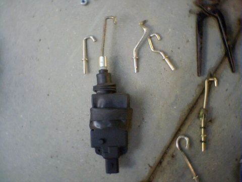 1994 Ford Bronco - A black car door lock actuator and various metal linkage rods are laid out on a gray surface.