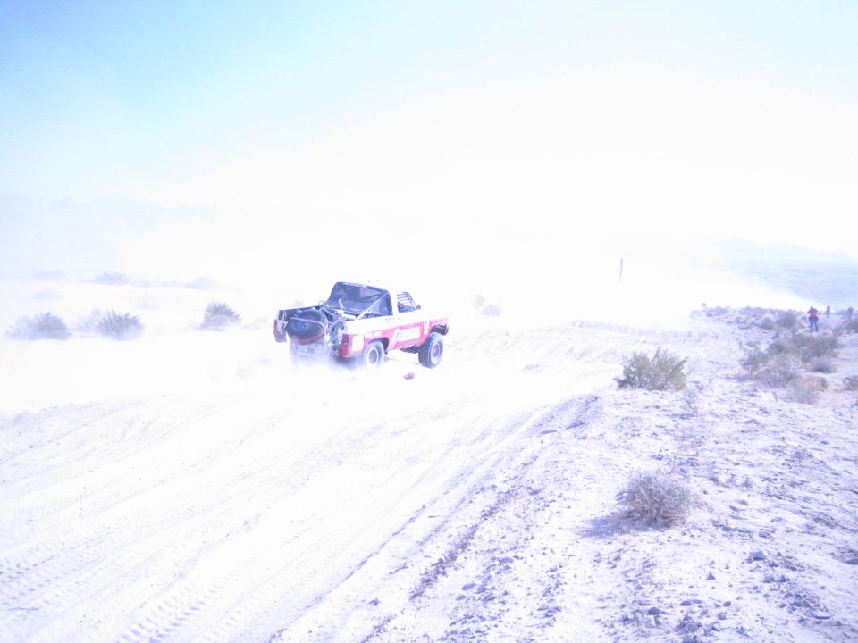 Red and white race truck with spare tire on a desert berm, spectators