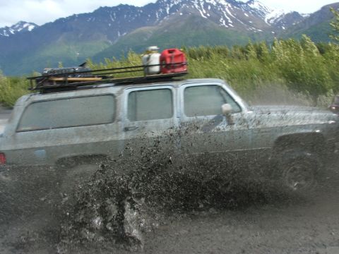 1990 Chevrolet Suburban - A 1990 Chevrolet Suburban kicks up mud as it drives through a muddy terrain with mountains in the background.