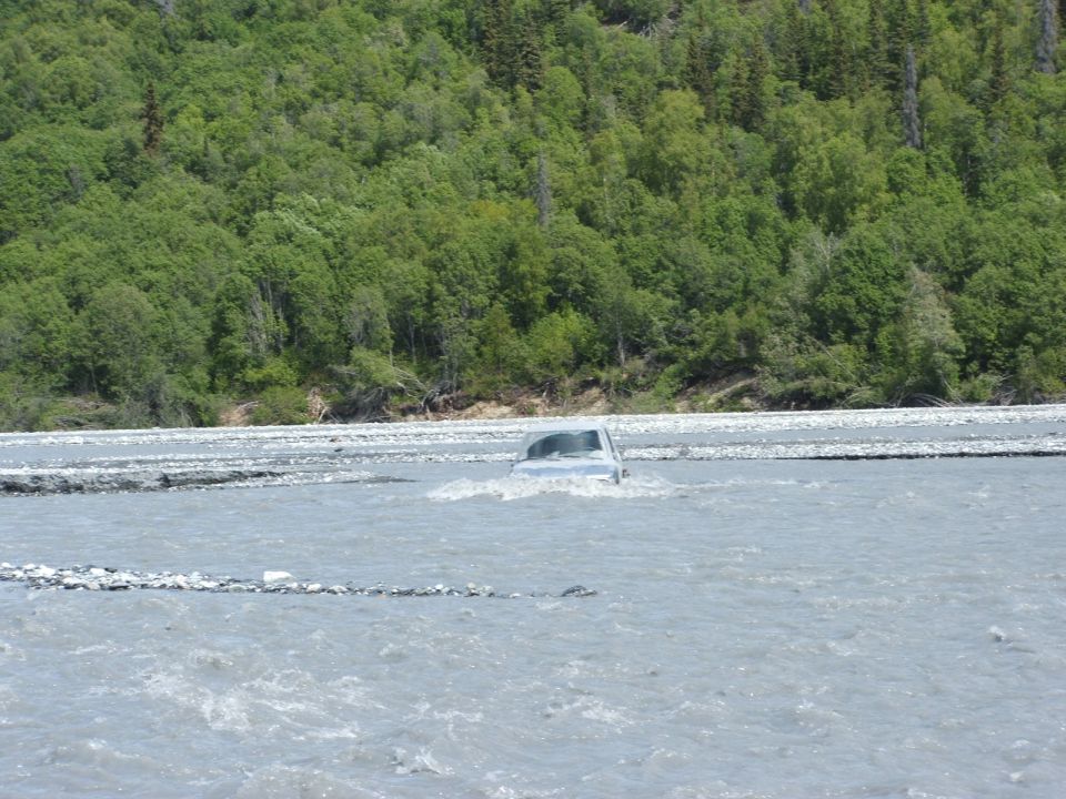 Silver vehicle deeply submerged while crossing a wide gray glacial river, forest in background