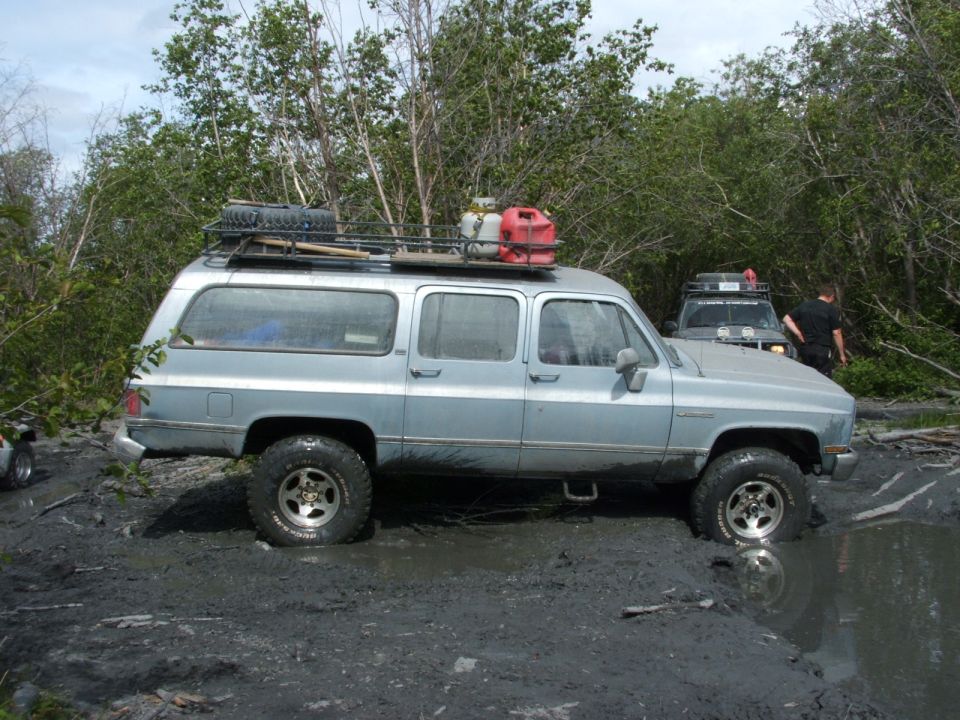 Lifted silver Suburban with loaded roof rack sitting in deep mud on a forest trail, second truck behind