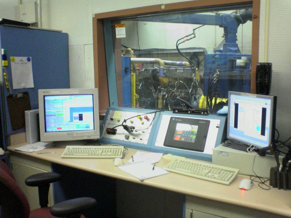 Engine on a test stand viewed through a window, with control consoles and monitors in the foreground