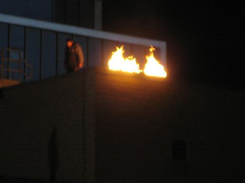 1990 Ford Bronco - At night, flames burn along the edge of a building roof while a person stands on a railed walkway nearby.