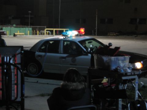 1990 Ford Bronco - police car with emergency lights flashing sits at night near stacked equipment and a seated person in foreground.