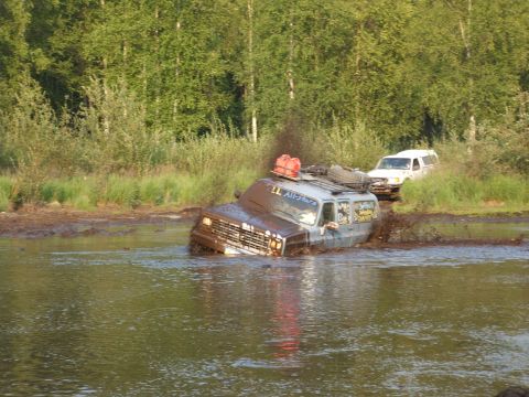 1990 Chevrolet Suburban - Two SUVs navigate a muddy water obstacle during an off-road excursion.
