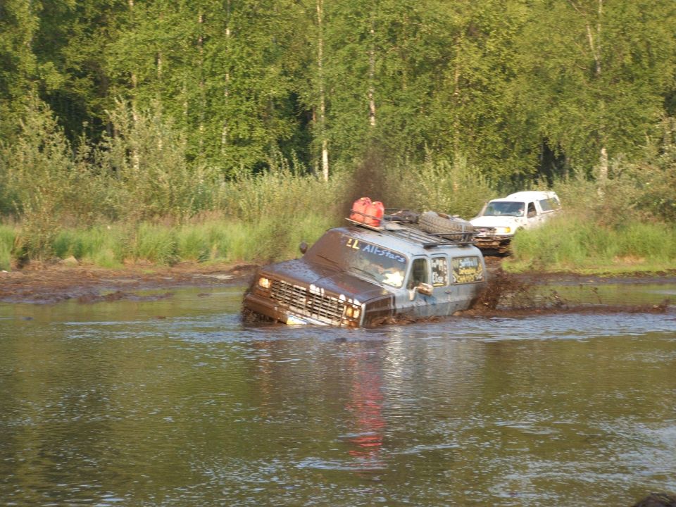 Square-body GM SUV with roof rack fording a deep muddy water hole off-road