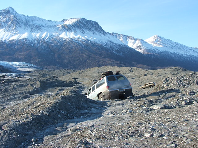 Silver SUV with roof rack crossing a rocky glacial moraine below snowy peaks
