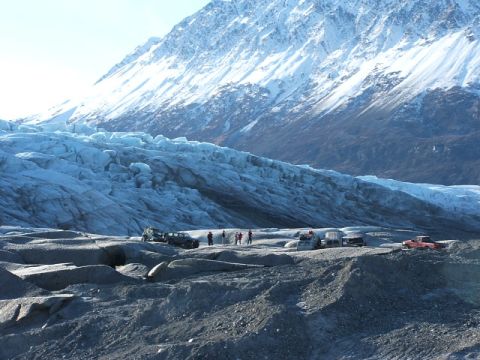 1990 Chevrolet Suburban - Multiple vehicles are parked on a rocky moraine in front of a vast glacier and a snow-covered mountain range.