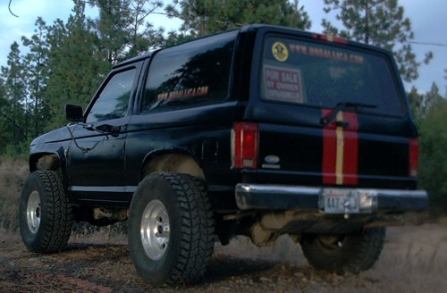 Black lifted Ford Bronco II with red stripes and For Sale sign, rear three-quarter view on a dirt road among pines