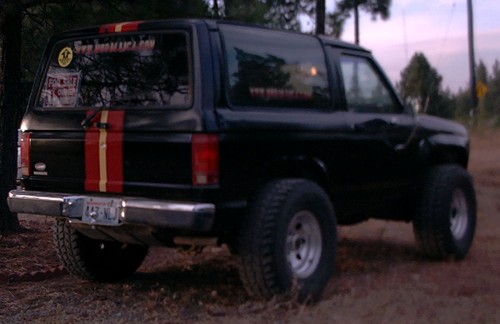Black Ford Bronco II with red/yellow stripes, lifted on big tires, rear three-quarter view among pines at dusk