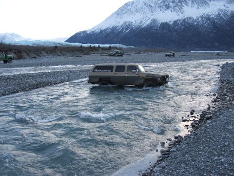 1990 Chevrolet Suburban - A modified Chevrolet Suburban navigates a rocky riverbed with a glacier and snow-capped mountains in the distance.