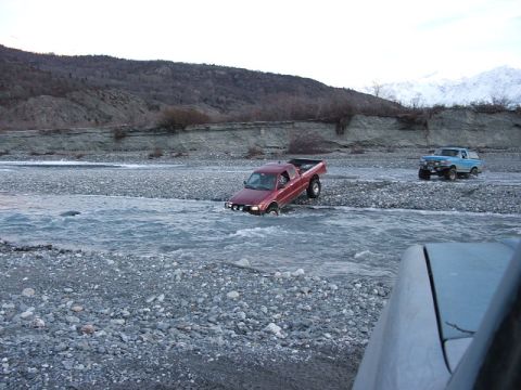 1990 Chevrolet Suburban - Two trucks navigate a shallow, rocky river with mountains in the distance.